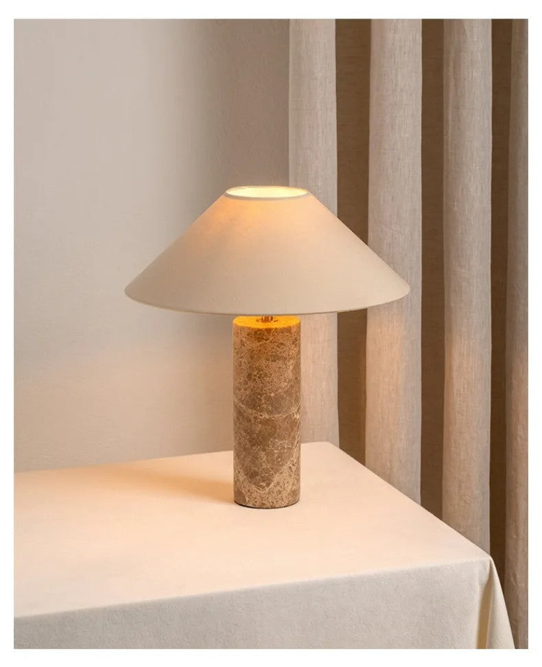 Table lamp with a textured base and beige shade on a tablecloth-covered table.